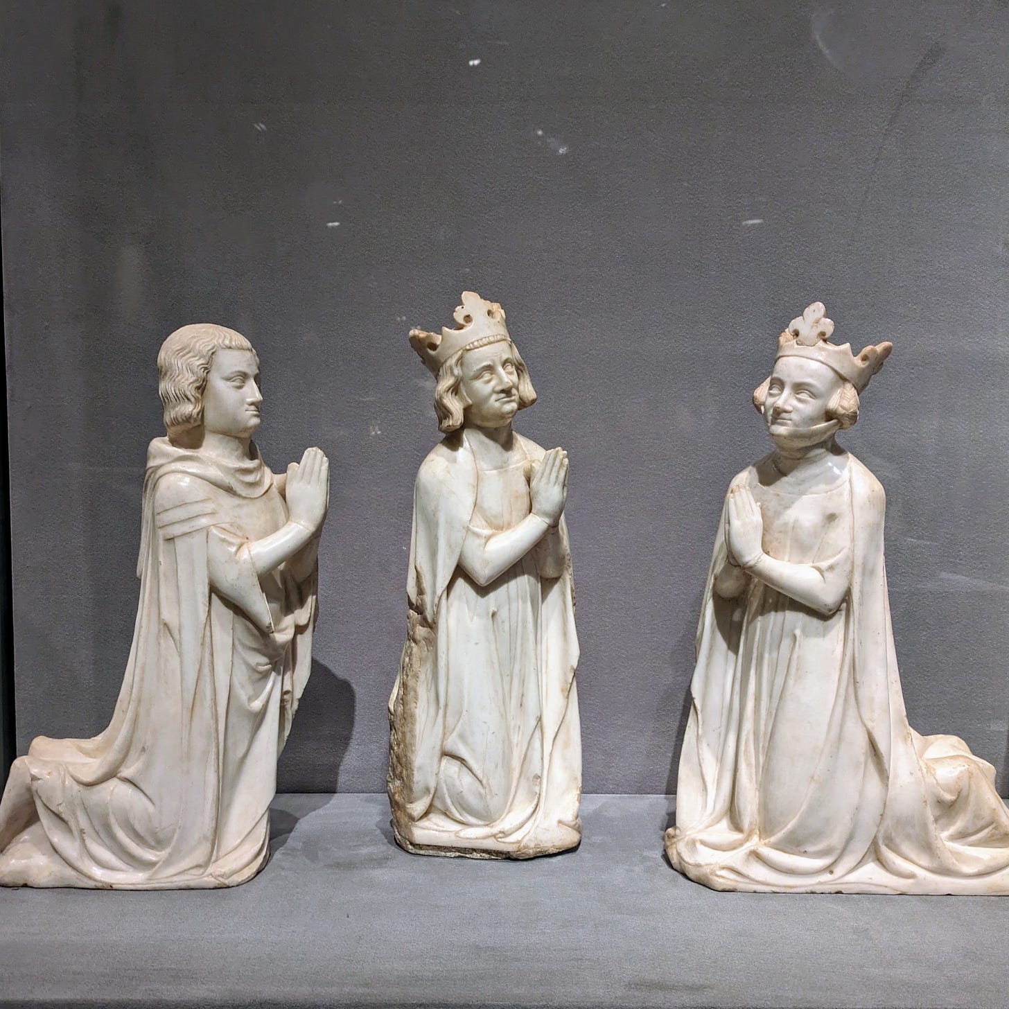 Photo of a museum vitrine, lined with grey felt, with three kneeling figures in yellowed white marble, hand joined in prayer chest-height, from left to right: a young man with chin-length wavy hair, a king with chin-length wavy hair, both looking to the right of the picture, and a queen with two ear-height buns of braided hair, looking to the left.