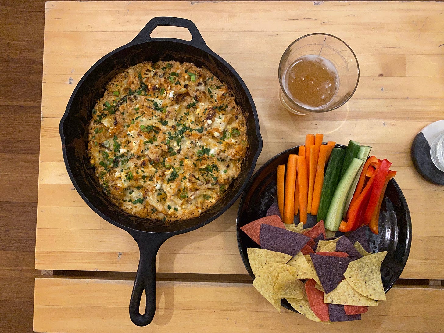 On the coffee table, a cast iron pan of creamy orange baked cauliflower dip, with sprinkles of parsley on top. Next to it is a glass of pale ale, and a black bowl with tri-colour tortilla chips, carrot and cucumber sticks, and red pepper slices.