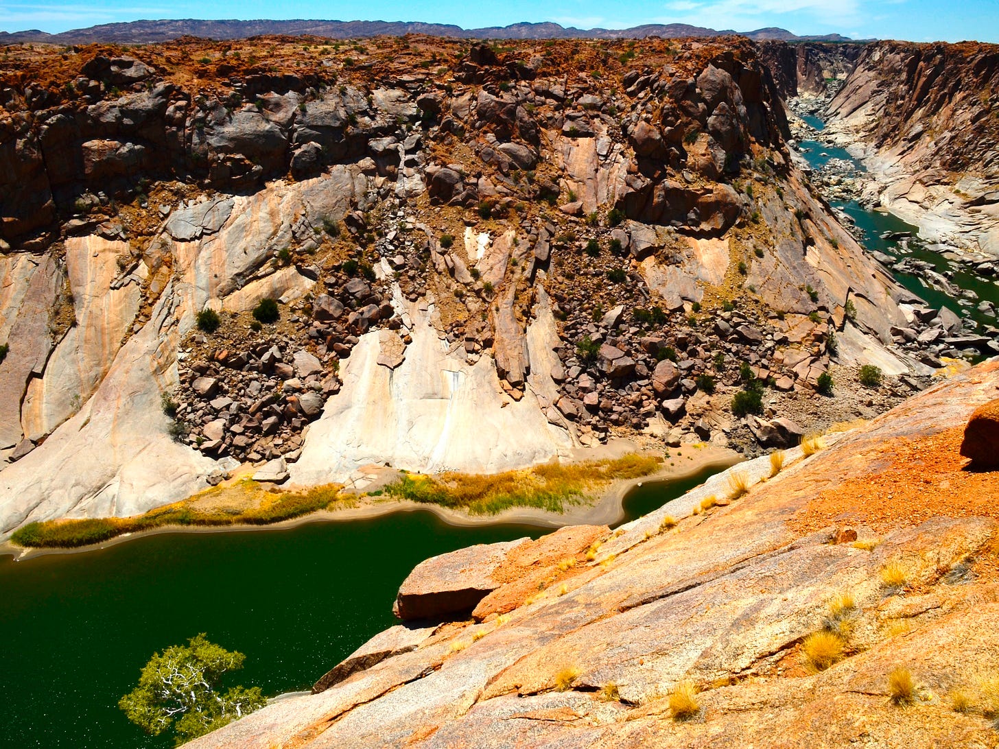 A wide canyon landscape under bright sunlight, with layered rock formations, steep cliffs, and a green river winding through the valley—captured from a high vantage point looking across and down into the canyon.