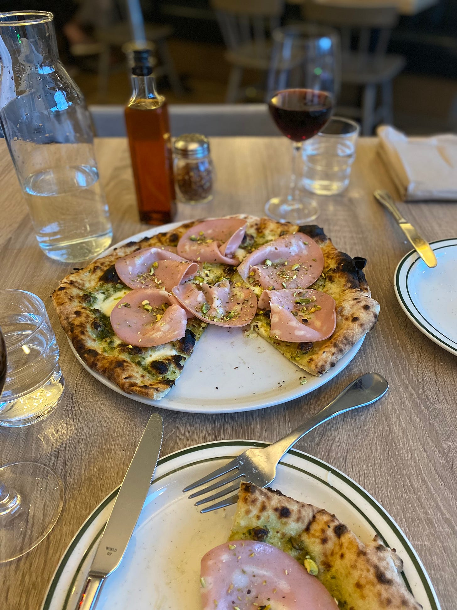 A table with drinks and condiments scattered around a large plate of the mortadella pizza above. The crust is browned and bubbled from the oven, and pesto and cheese are visible beneath slices of mortadella arranged on top with chopped pistachios over them. One slice is missing from the pizza, and can be seen partially on the plate in the foregrount, which has a knife and fork resting at angles on the edge.