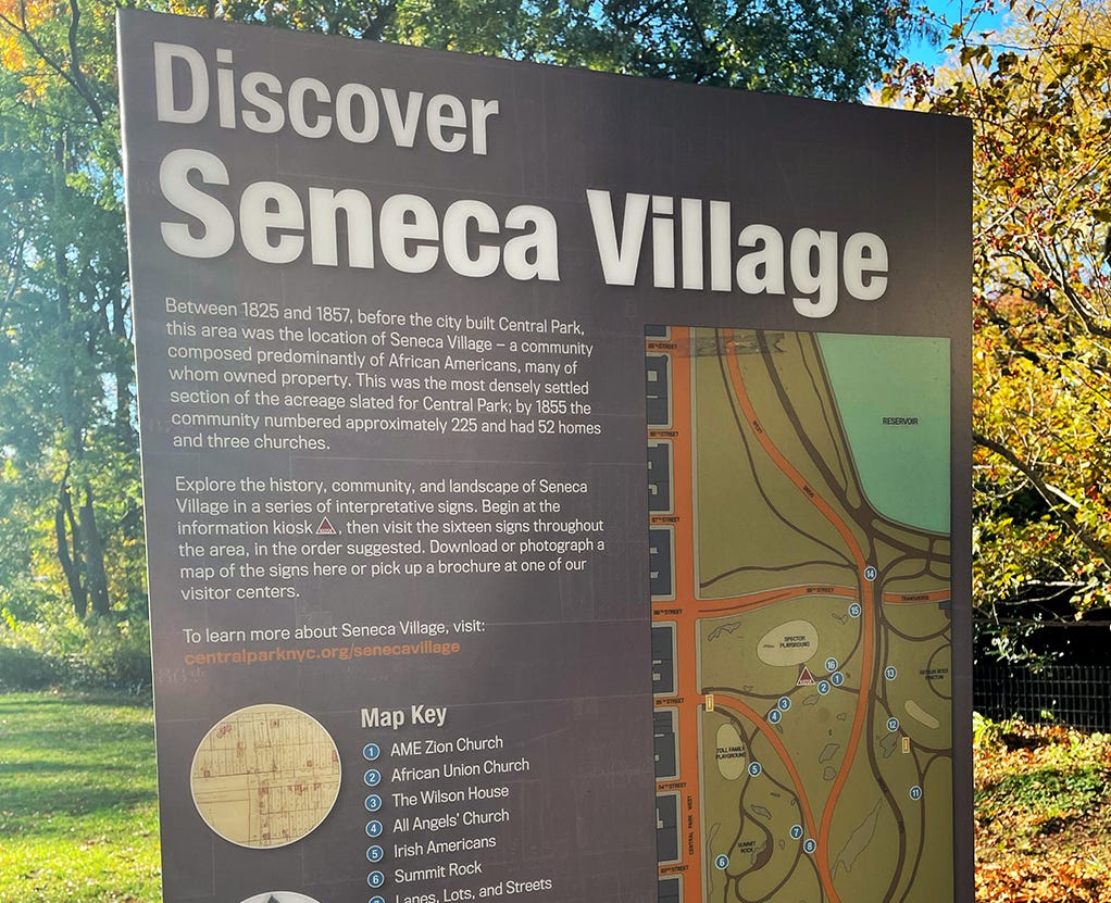 A photograph of a sign in Central Park that says "Discover Seneca Village" and describes the historical site.