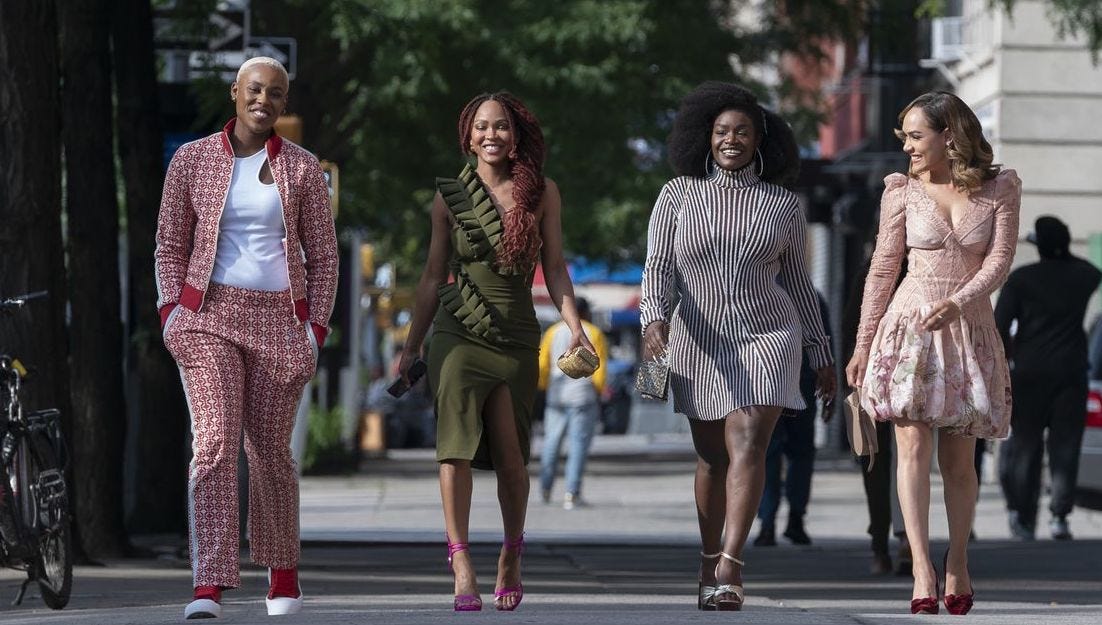 Four black women walk side-by-side toward the viewer. They are all very different types of women, though all impeccably dressed in distinct styles. They are all smiling as they walk together on the sun-dappled city sidewalk.