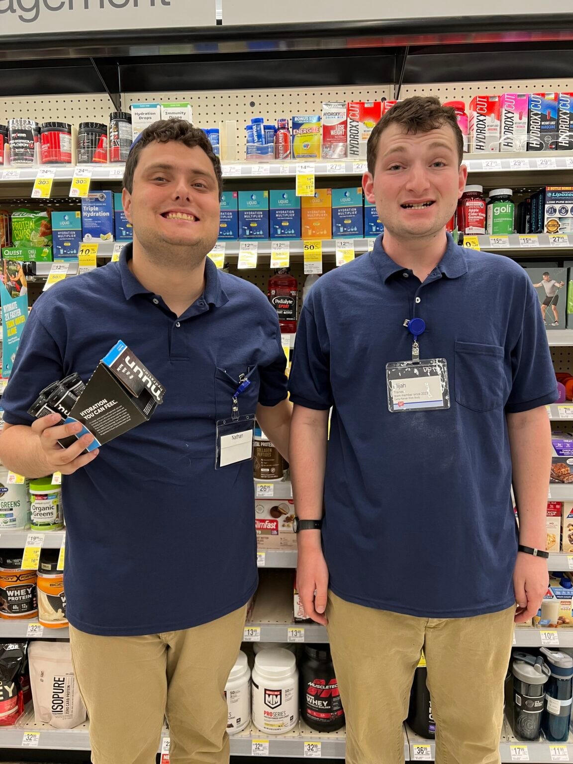 Two young men in polo shirts and wearing work IDs stand in front of a pharmacy shelf