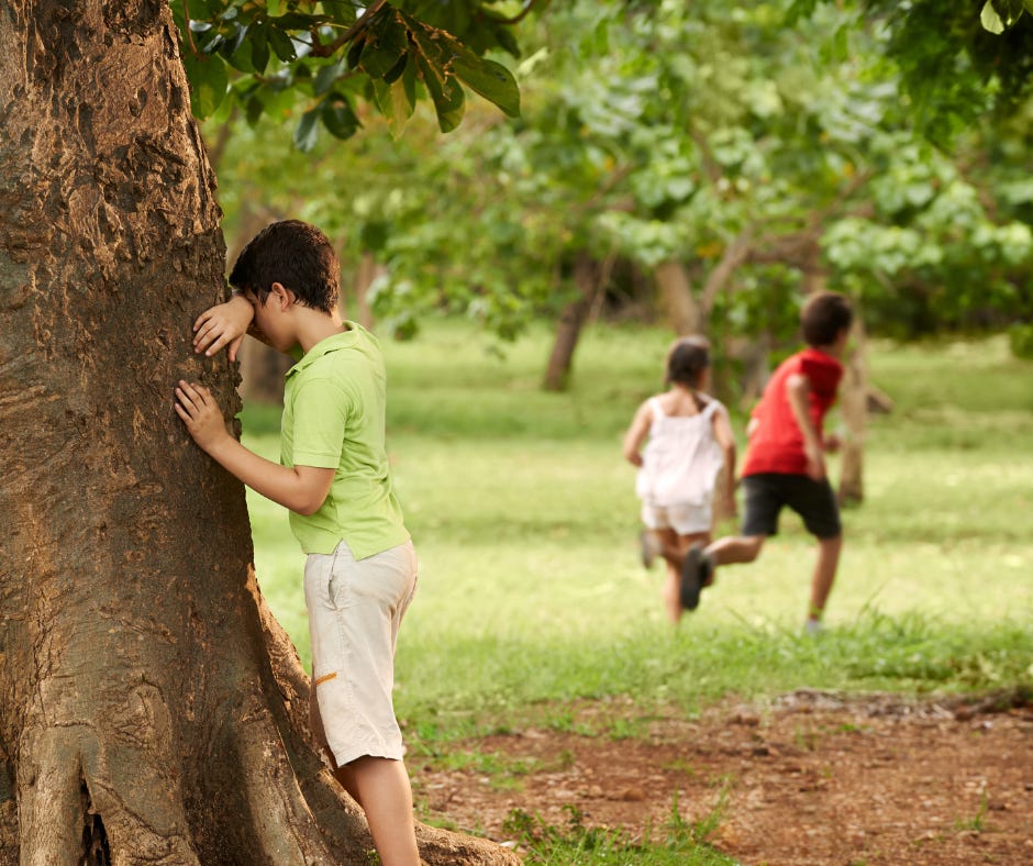 two children run into the trees in the distance while a third child leans up against the trunk of a tree in the foreground, burying his face against his arm so he can't see