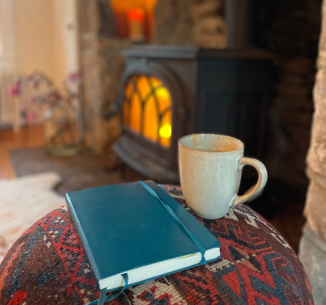 In the foreground, a cup of coffee and a journal with a teal green cover rest on an ottoman covered in a North African woven tapestry. In the background is a hearth with a fire glowing in a wood stove. 