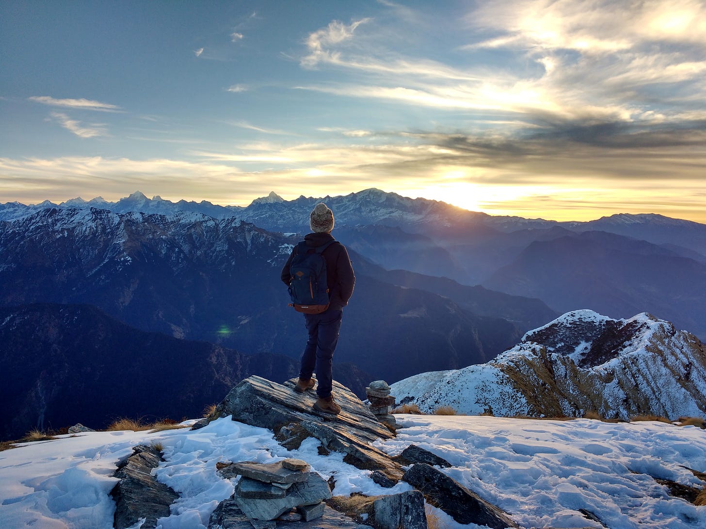 A person standing on a rock overlooking a snowy mountain range at sunrise. The person is waering a white knit hat, a black jacket and black pants with brown boots. They are also wearing a blue backpack.