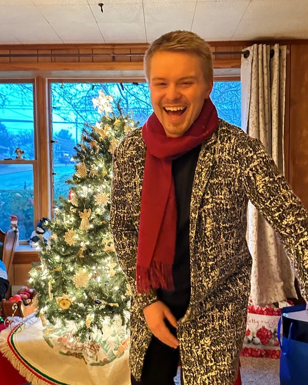 Young White man with goatee, in black/white  long jacket and deep red scarf, laughing in front of small, lit Christmas tree and picture window, indoors.