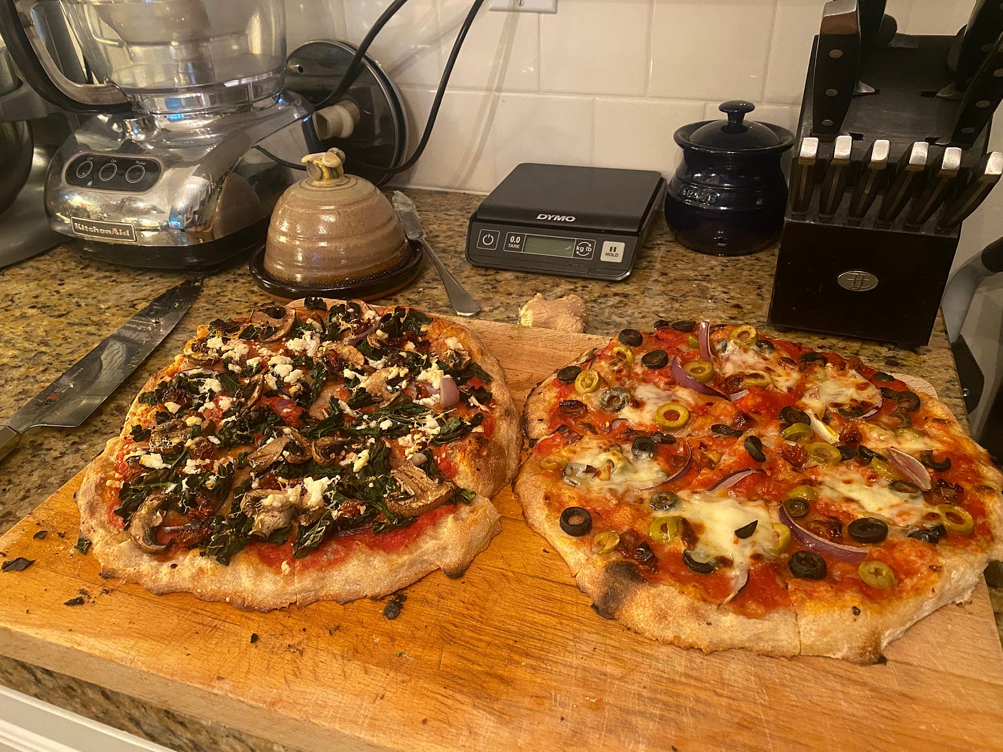 Two pizzas on a cutting board, the one on the left with kale as described above, and the one on the right with olives and fresh mozza. In the background are a butter dish, a kitchen scale, and a knife block. A large knife rests on the counter beside the cutting board.