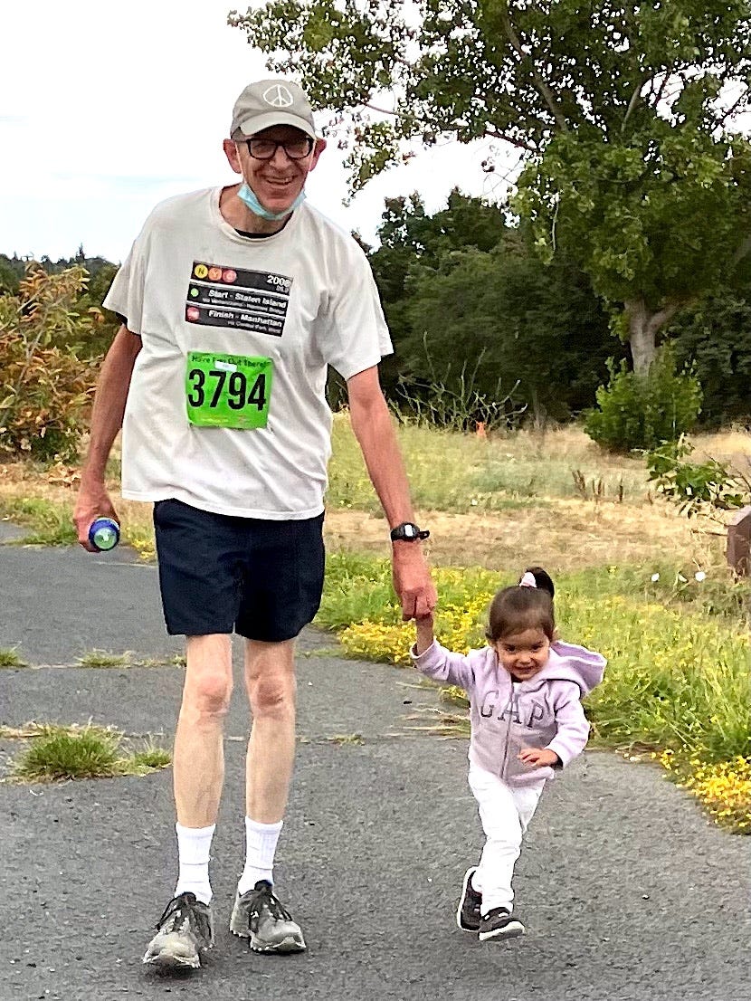 Photo of a tall pale skin man, smiling big, wearing a runners outfit, on a road with trees and grass alongside it. He is holding the hand of a young girl, maybe 2-3 years old, who is pulling him along, as she runs in front of him.