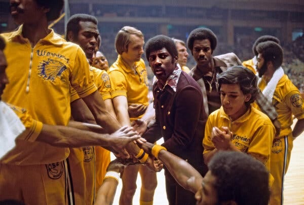 Attles, wearing a brown suit, stands on a basketball court as several Warriors players, in yellow uniforms, surround him.