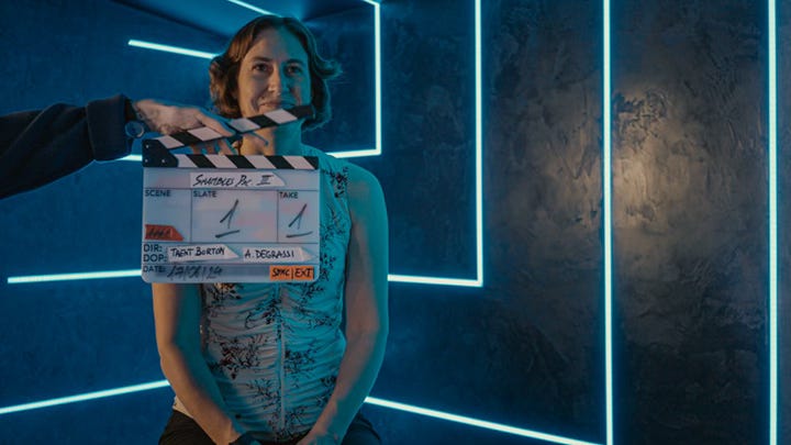 A woman in a white top sits on a stool in a studio with a clapperboard in front of her that reads 'Shambles Doc III" Take 1'