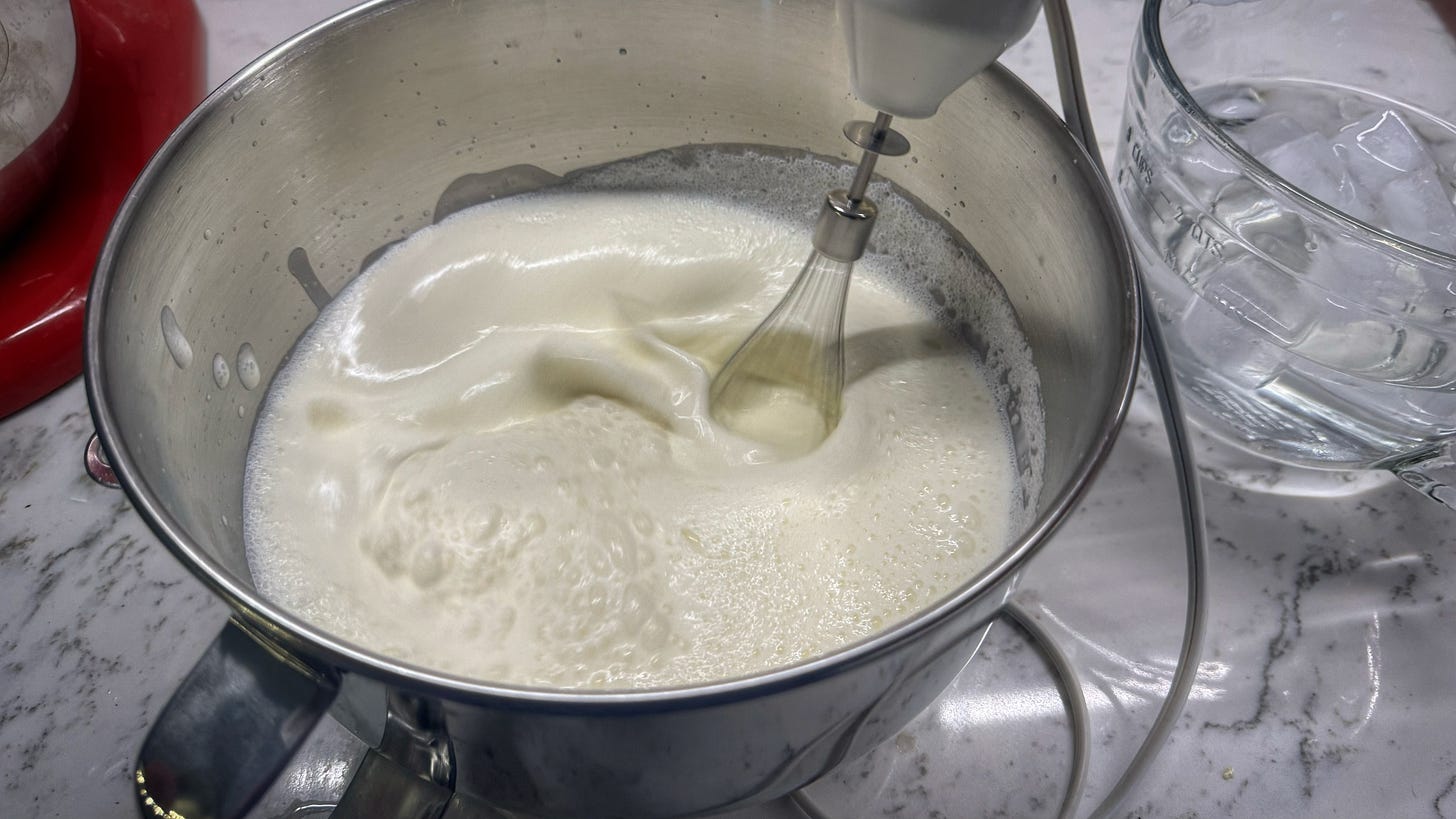 The whisk attachment of an immersion blender splashing through cream in a steel mixing bowl. There is a measuring cup full of water and ice cubes beside it. 