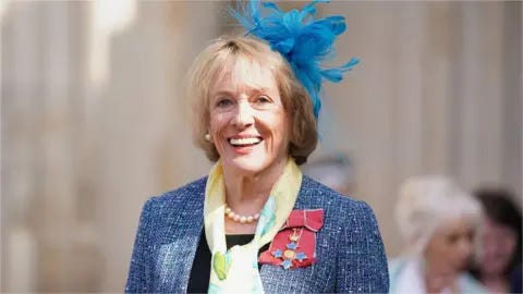 PA Media Dame Esther Rantzen smiles at the camera. She has a blue-feathered fascinator in her shoulder-length blond hair. She is wearing a blue suit and yellow scarf with a medal on a pink ribbon, a pearl necklace and pearl earrings.