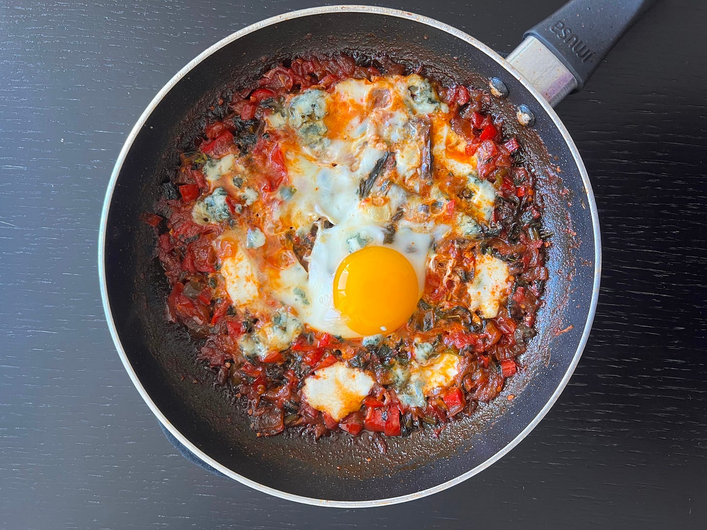A homemade shakshuka in a black frying pan, featuring a rich, deep red tomato sauce with melted blue cheese and a perfectly set egg in the center. The dish is slightly charred at the edges, enhancing its rustic look. This shakshuka is part of Liat Portal’s Foodie Disorder, where she explores flavors, textures, and the art of cooking.