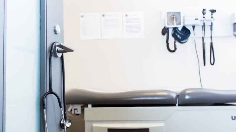 A doctor's examination room is seen through an open door, with a stethoscope hanging on its handle.