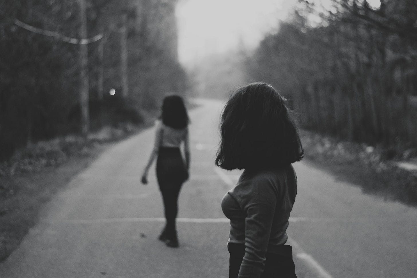 black and white image where one woman watches the other behind her back while both stand on an empty country road