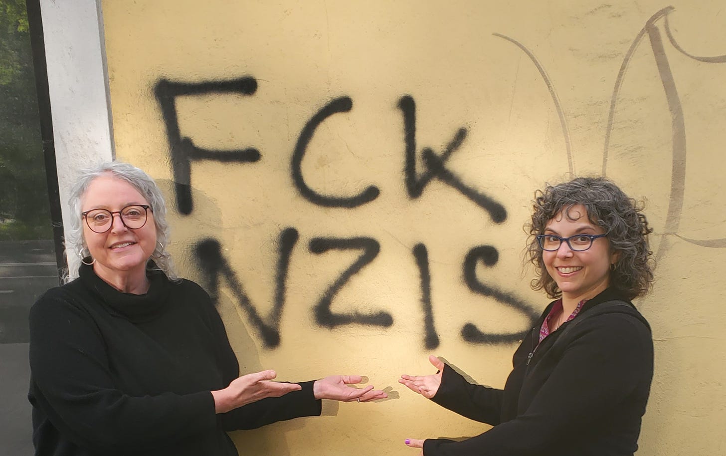 Two Jewish women in black tops, each wearing glasses, stand in front of graffiti that reads FCK NZIS