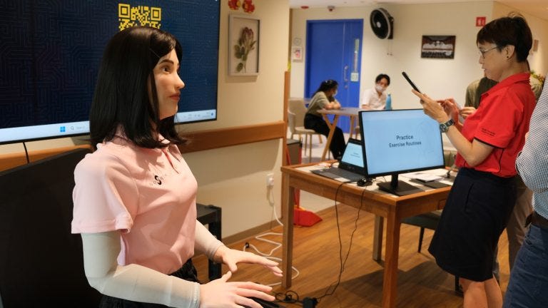 A humanoid robot with long black hair is seated in a nursing home setting, wearing a pink shirt. In the background, a woman in a red shirt is using a smartphone, while a computer screen displays 