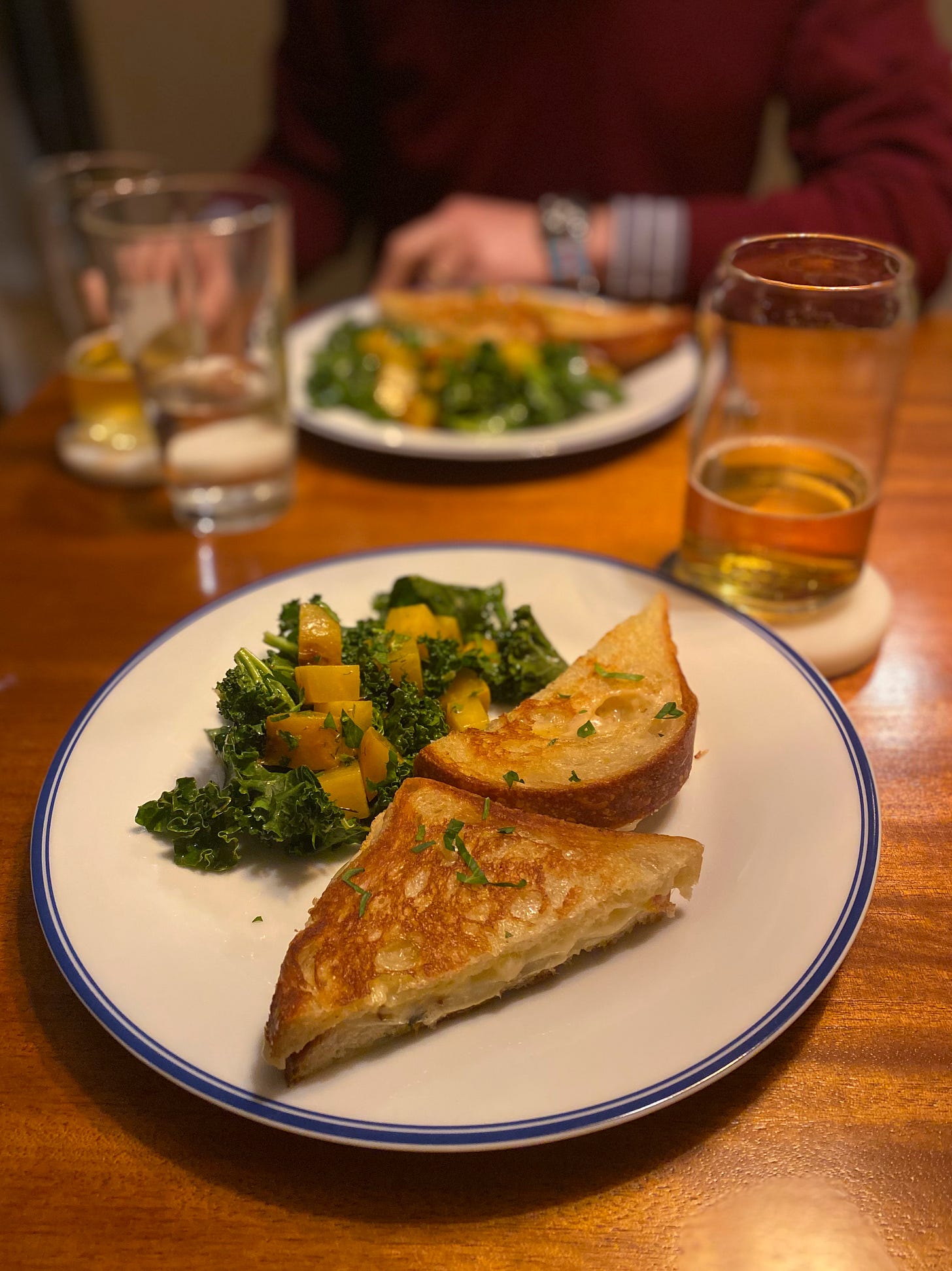 A halved grilled cheese sandwich with roasted fennel slices next to a kale and golden beet salad on a white plate with a blue rim. Next to it is a partially full glass of beer on a coaster. Jeff sits across the table, blurred in the background, the same meal in front of him.