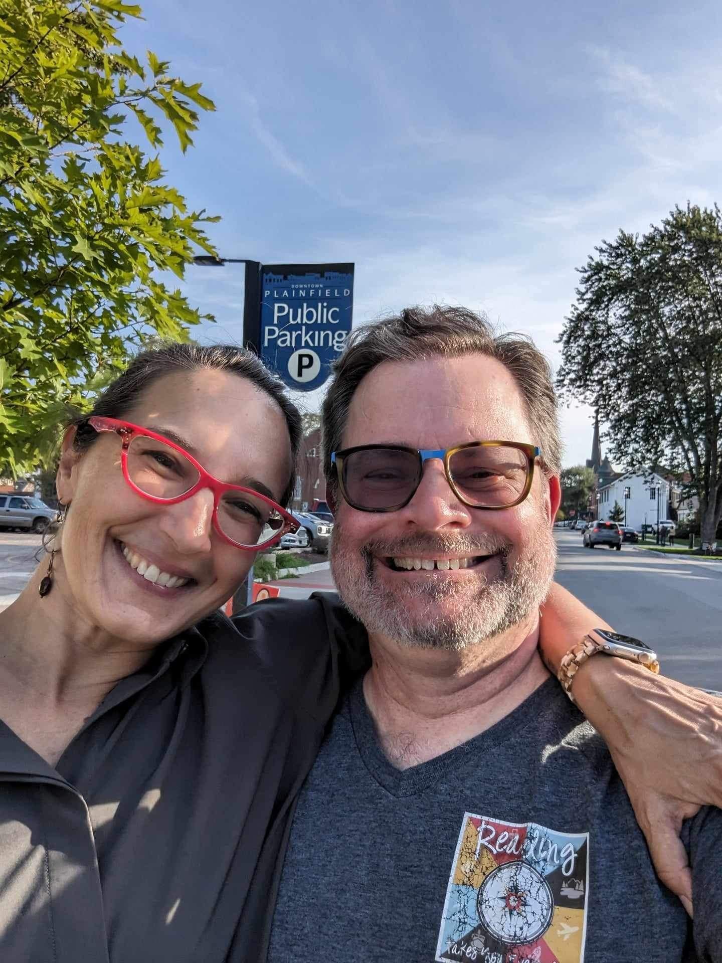 Sonya smiling with red glasses, arm around Carl, smiling with polarized glasses, trees and the library and blue sky in background and a sign hanging from the lamp post that says Plainfield Public Parking