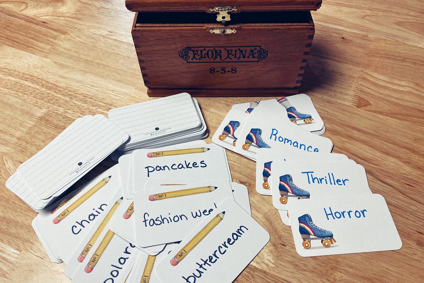 A bunch of cards with words written on them, spread out on a light wood table, with a cherry wood cigar box cracked open behind them. The cards on the left read "pancakes, fashion week, buttercream" with a few more only partially visable and two stacks of cards behind them. The cards on the right read "romance, thriller, horror" with a few more piled underneath them.