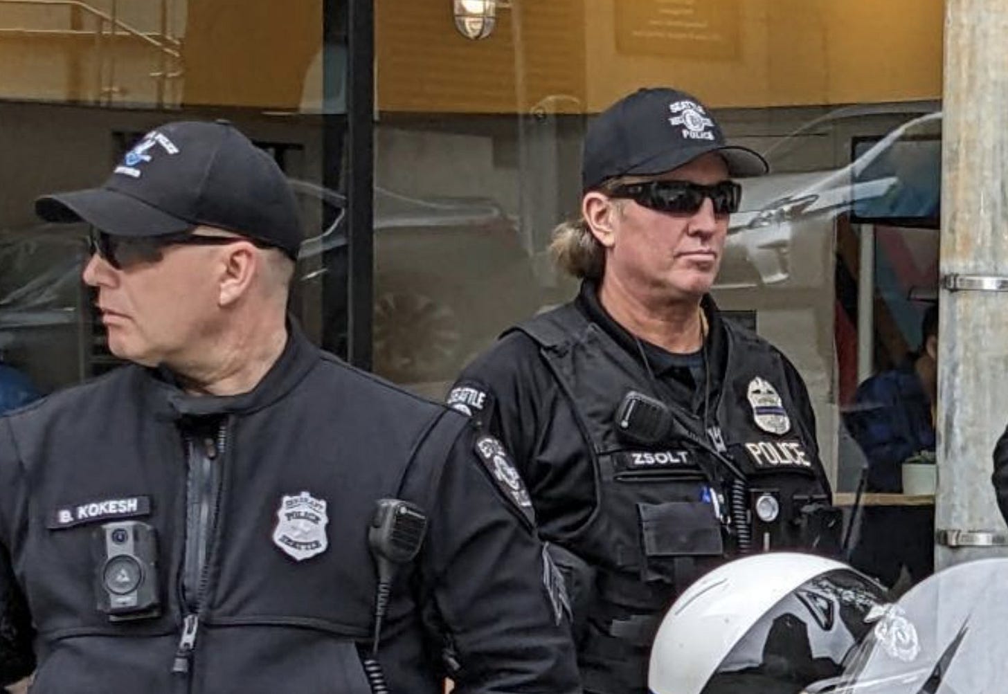 Two Seatle police officers in their 50s wearing black ballcaps and sunglasses looking in opposite directions. 