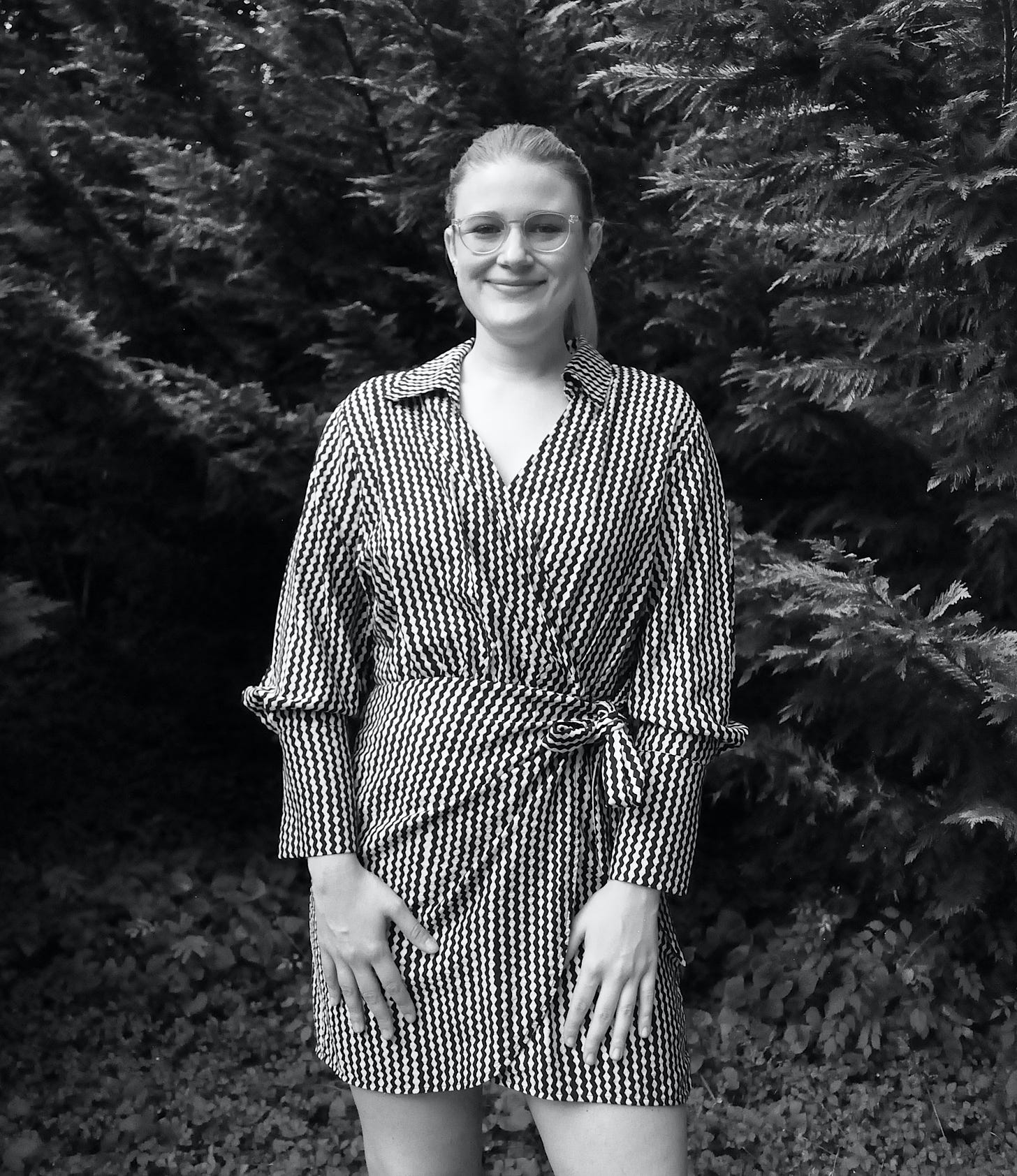 Photo of Shady Kimzey, a white non-binary femme wearing a mid-legnth black and white wrap dress, wearing glasses with their hair in a ponytail, standing in front of evergreen trees outside.