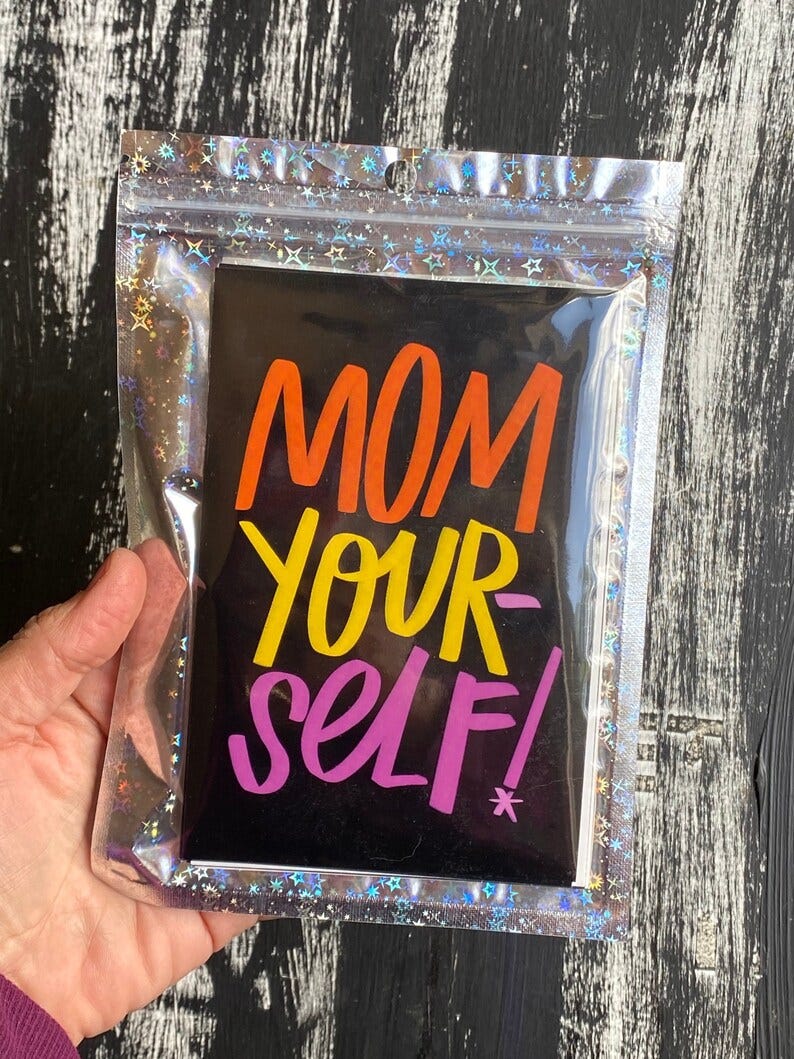 May include: A black card with the words 'MOM YOUR SELF!' written in orange, yellow, and pink lettering. The card is in a clear plastic bag with a holographic pattern of stars.