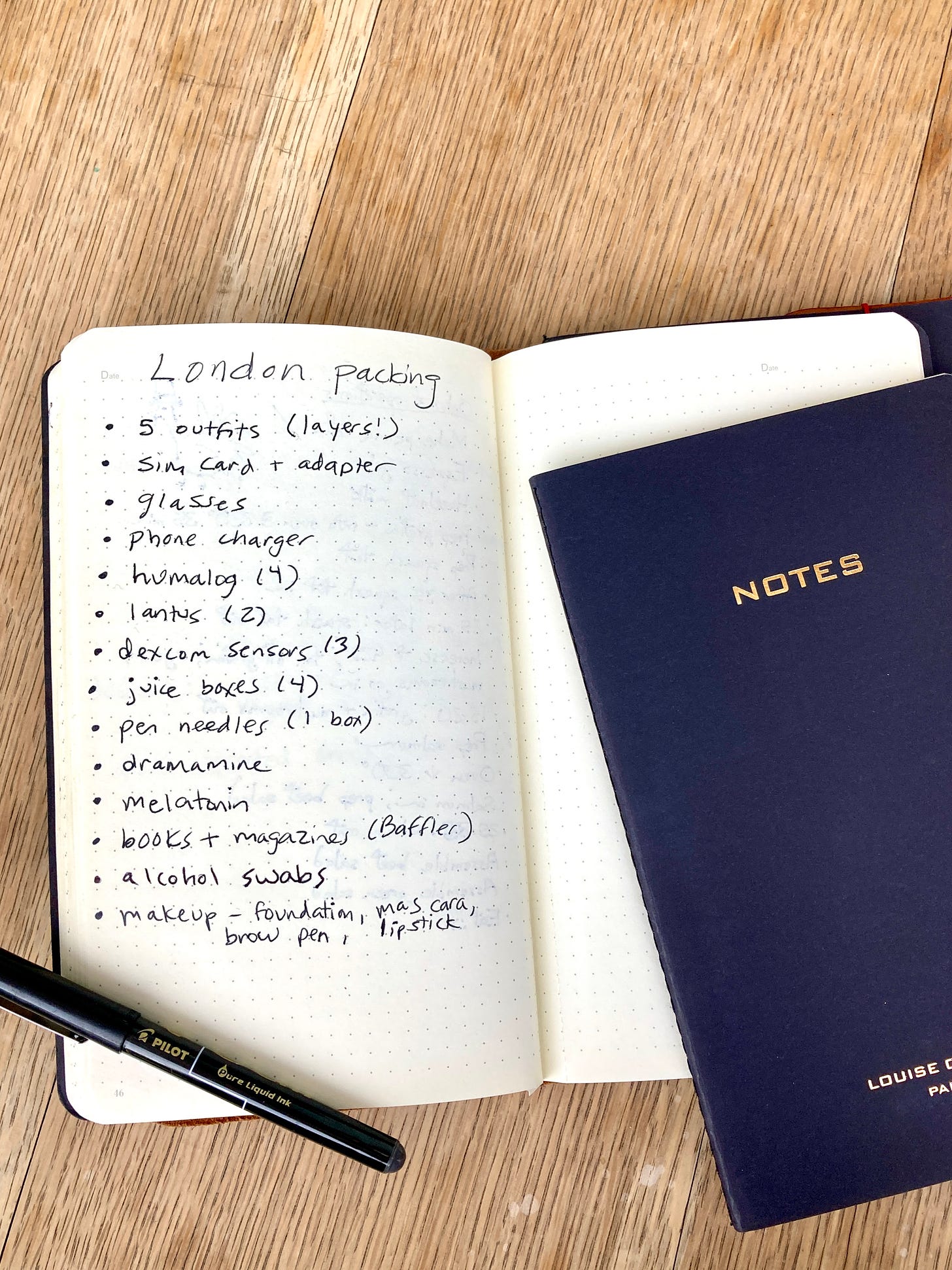 A notebook on an textured wood table. It is open and the left page is titled "London Packing." The page has items listed in bullets, including clothing, medicine, make-up, and other essentials.