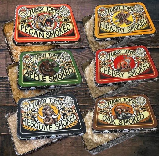 Six tins that look like tobacco tins all in different colours: red, orange, black, green, brown and black, with tattoo-inspired artwork. Each tin reads "Tubby Tom's Gloucester" and the type of salt inside.