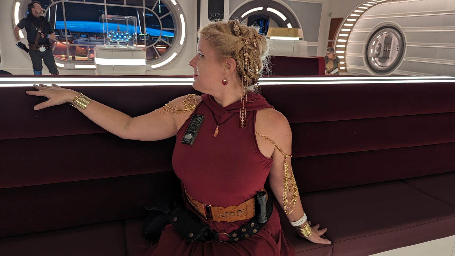 A pale-skinned blonde woman sitting on a long red couch on the Halcyon Starcruiser, with the ship's bridge visible in the background. Her hair is elaborately styled with coils, braids, and dangling gold jewelry. She wears a red hooded sleeveless dress with a combadge on its chest, draping golden shoulder jewelry, a light brown belt with a fuel canister attached, golden bracelets, and a black hip pouch belt. She is turned partly away from the camera, looking at something off to her right, one hand resting on the back of the couch.
