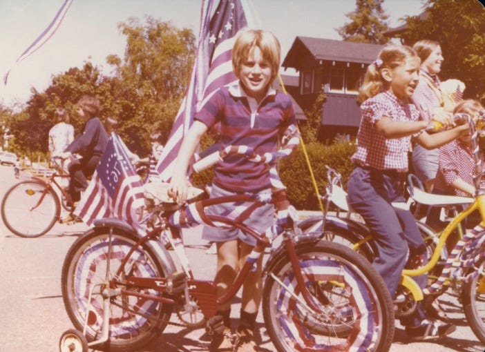 A blonde five-year-old boy posing with a bike decorated with red, white, and blue bunting with bicentennial US flags in the background.