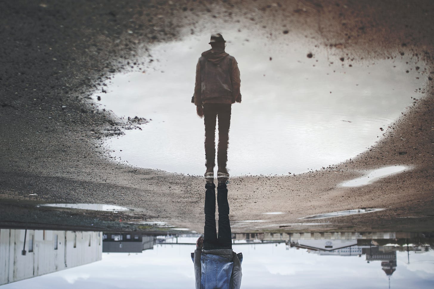A gray puddle with a reflection of a man wearing a jacket, jeans and a fedora. 