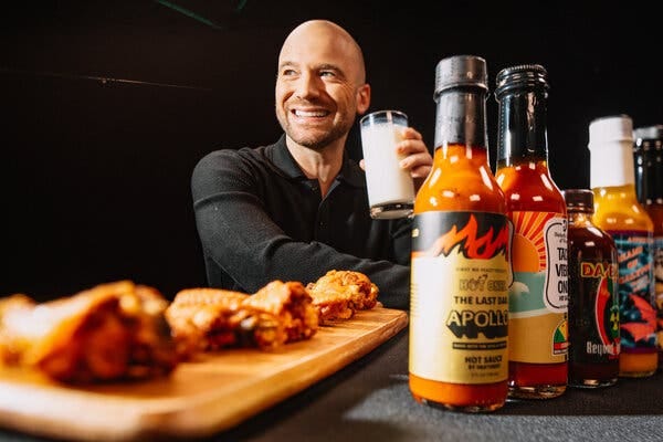 A man sits holding a glass of milk behind a table that has on it a wooden board with chicken wings and a row of hot sauce bottles. He’s looking to his right with a big smile. 