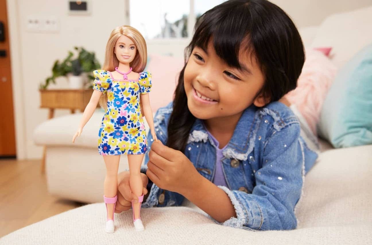 A young femme of color is playing with a Barbie with Down Syndrome who has blonde hair, wearing a floral dress and pink ankle foot orthoses..