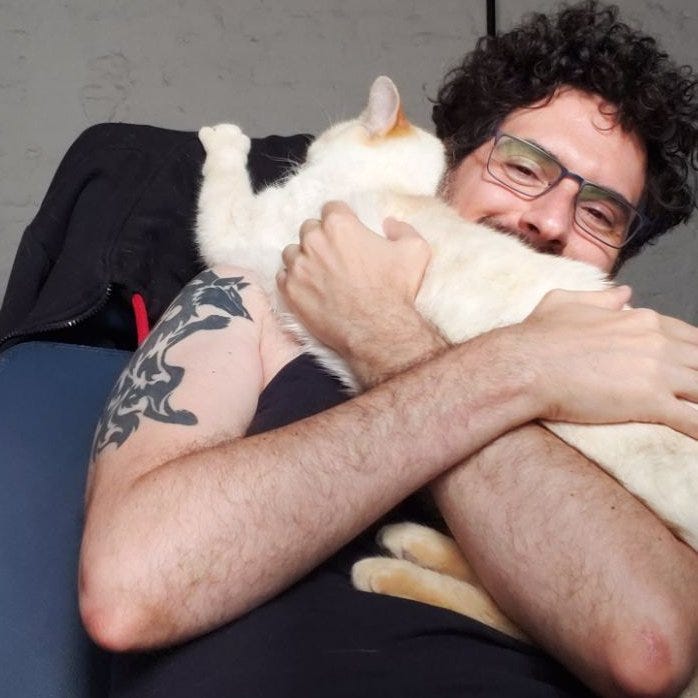 Imagem colorida: gato Dexter atravessado no meu colo, da barriga ao peito, segurando-se no encosto da cadeira do escritório, cabeça apoiada no meu pescoço. Tira um cochilo enquanto recebe um abraço.