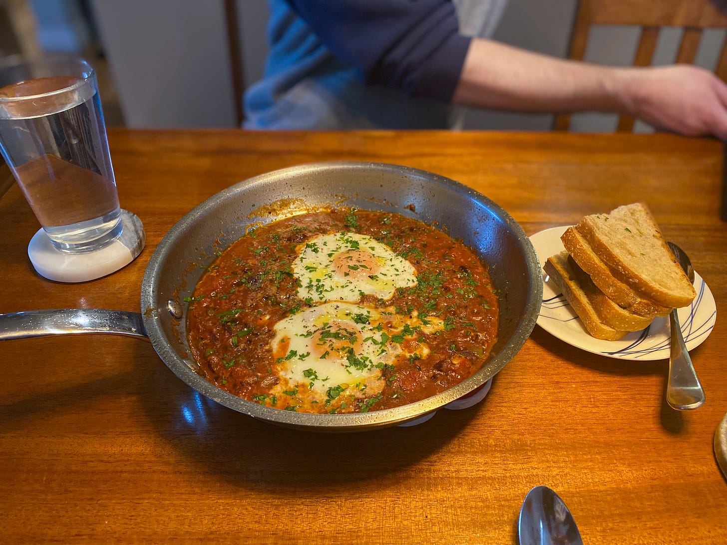 A table set with a stainless steel frying pan full of puttanesca, and topped with two poached eggs, parmesan, and parsley. Next to the pan is a small plate with slices of toast. Jeff sits across the table in the background.