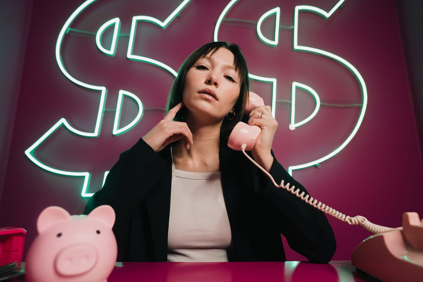 woman sitting in front of a neon dollar sign. She is holding a pink corded phone