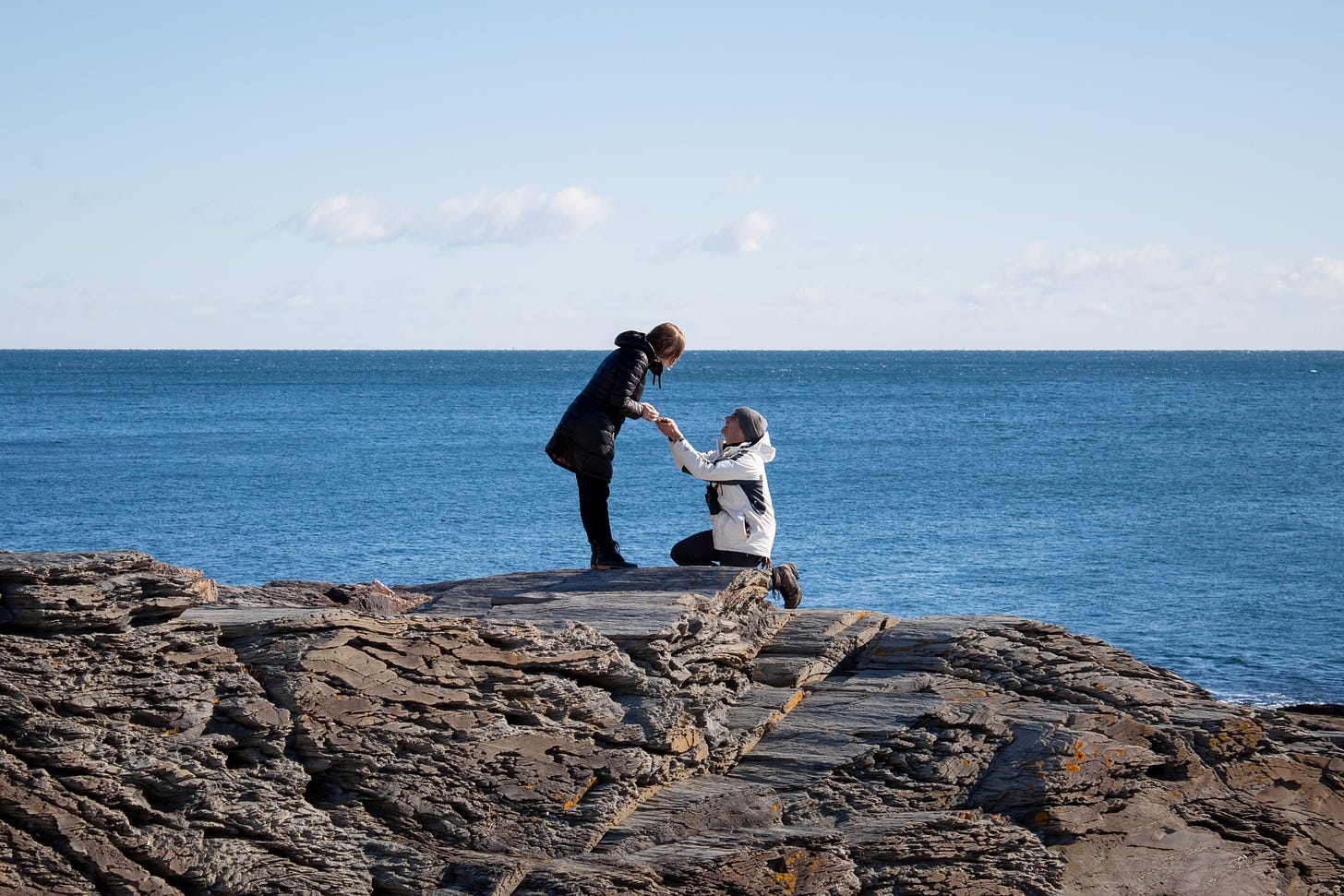 someone in a big white coat on one knee proposing to someone in a long black coat and black pants, on a cliff set against the ocean