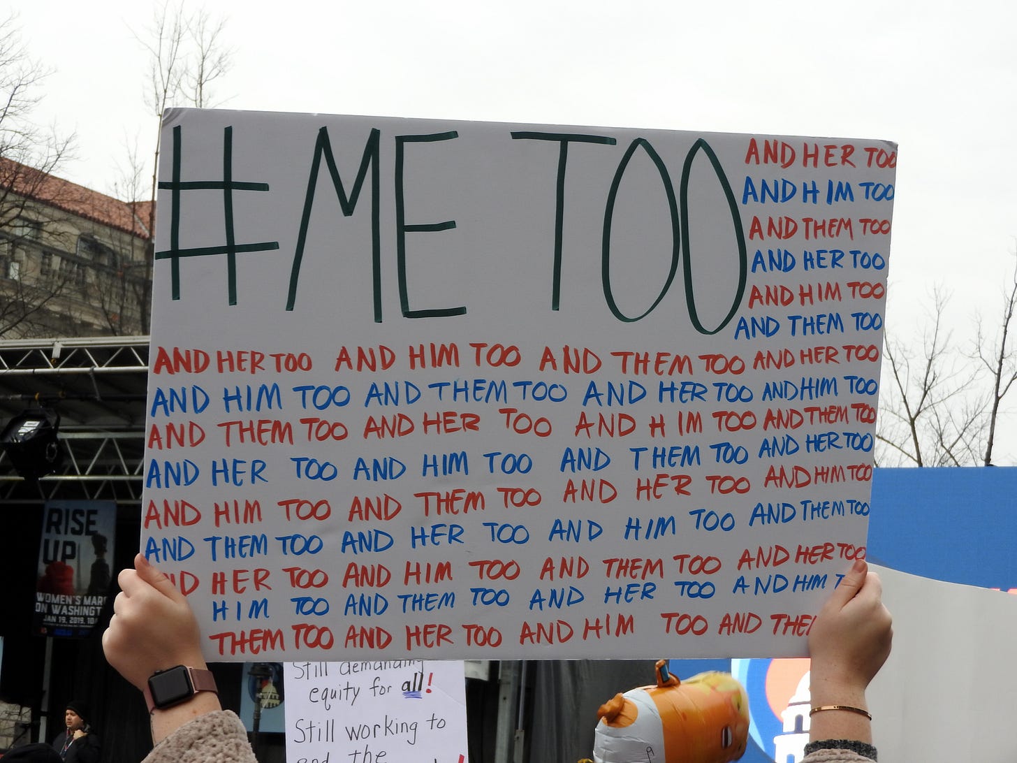 Person holding sign that says "me too and her too and him too and them too"