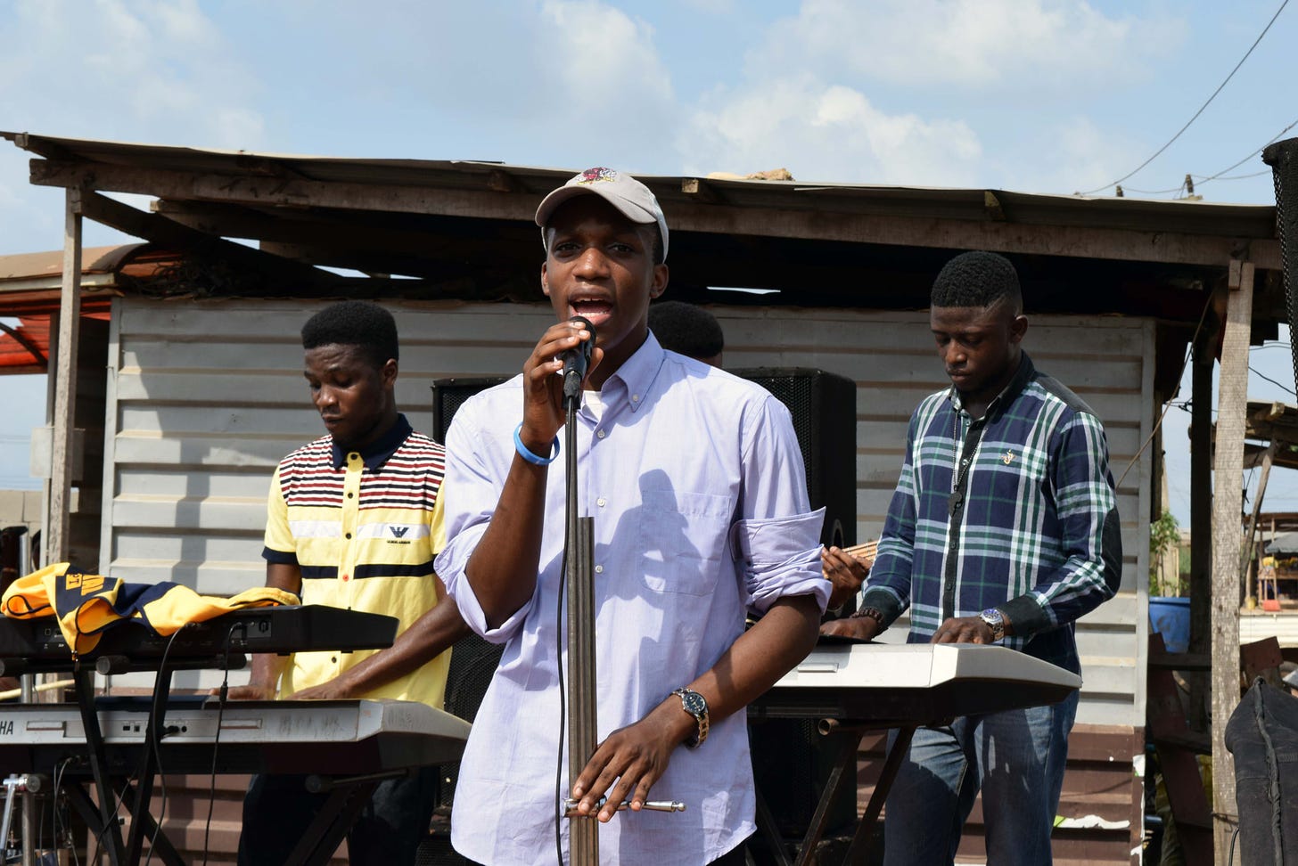 A picture of me outdoors, singing into a microphone on a mic stand. I am standing in front of two keyboardists, wearing a light purple shirt and a cap.