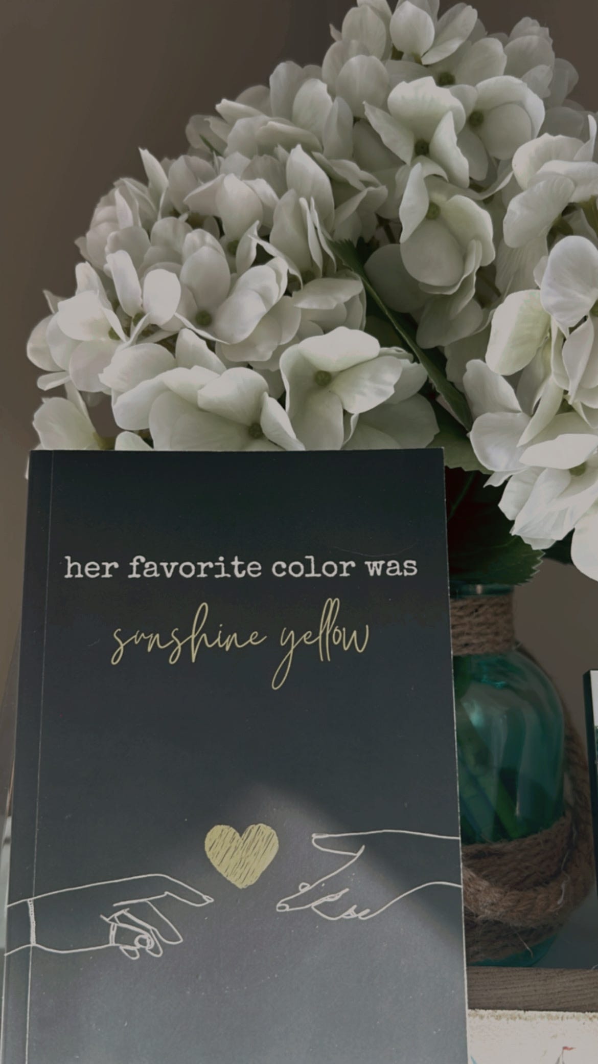 black cover of book with white line drawing of two hands outstretched towards each other with a yellow heart in the middle. title is "her favorite color was sunshine yellow." white hydrangea flowers are in the background in a blue-green vase with brown rope around it as accents.