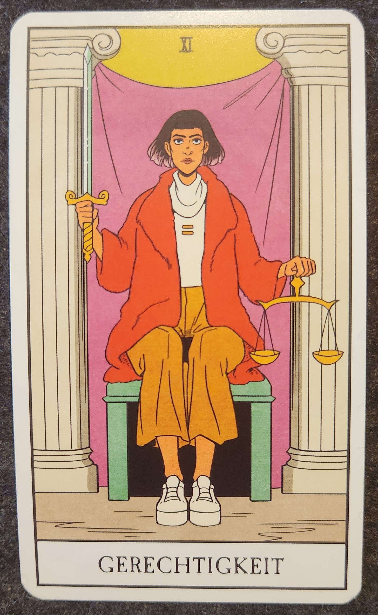 A tarot card labelled XI Gerechtigkeit. A person with a short brown bob is sitting unsmiling on a green bench between two ornate white pillars. There is a red curtain draped behind them. In one hand, they are holding a sword with the blade pointing up in the air. In the other hand, they are holding a set of balanced golden scales. 