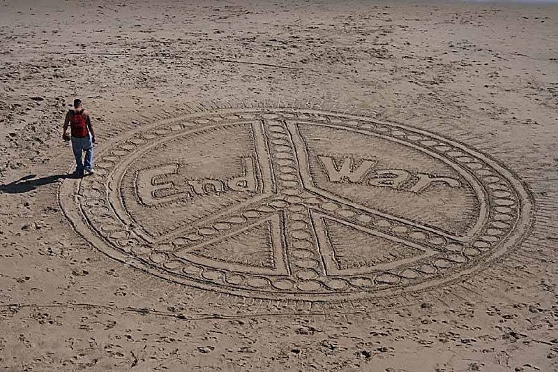 A lone person walking on the edges of a huge peace sign drawn in sand on the beach, with the words “End War” written inside.