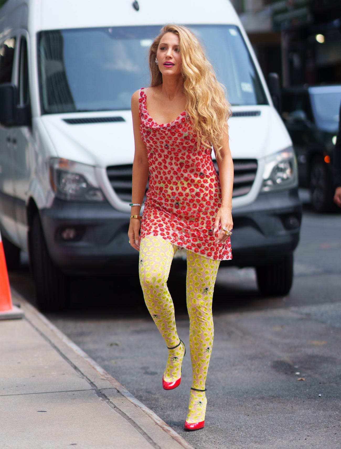 Blake Lively walks down the street in a red, floral dress with yellow tights and red shoes.