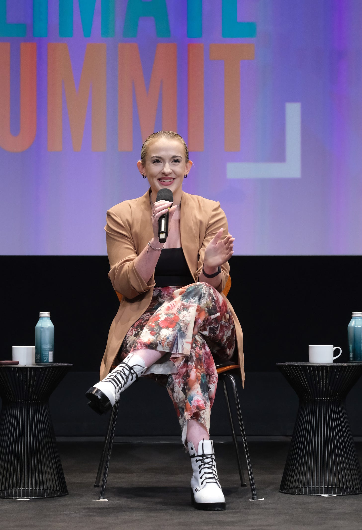 A blonde woman with slicked back hair is sitting on stage holding a microphone. She is wearing a camel-colored jacket, black tank top, and brightly patterned pants with white boots. Behind her is a purple digital screen that says "Climate Summit."