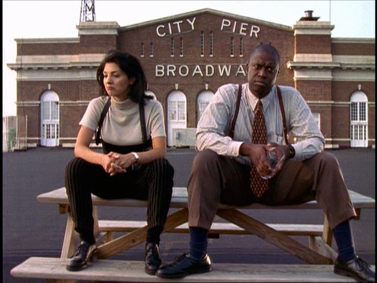 This still image from "Blood Ties (Part 3)" shows Detective Laura Ballard (played by Callie Thorne, left) and Detective Frank Pembleton (played by Andre Braugher, right) sitting atop a wooden picnic table on the Baltimore Homicide Unit building's roof. The words "City Pier--Broadway" are visible on a brick wall behind them. This still image from "Blood Ties (Part 3)" shows Detective Laura Ballard (played by Callie Thorne, left) and Detective Frank Pembleton (played by Andre Braugher, right) sitting atop a wooden picnic table on the Baltimore Homicide Unit building's roof. The words "City Pier--Broadway" are visible on a brick wall behind them.