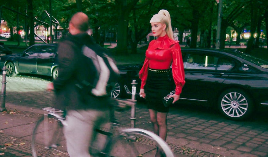 Erka standing on a sidewalk in Berlin. She is wearing a red latex turtle neck and a black latex mini skirt. Her blond hair is in a high pony tail with sharp bangs. A blurry man on a bicycle rides past, almost hitting her.