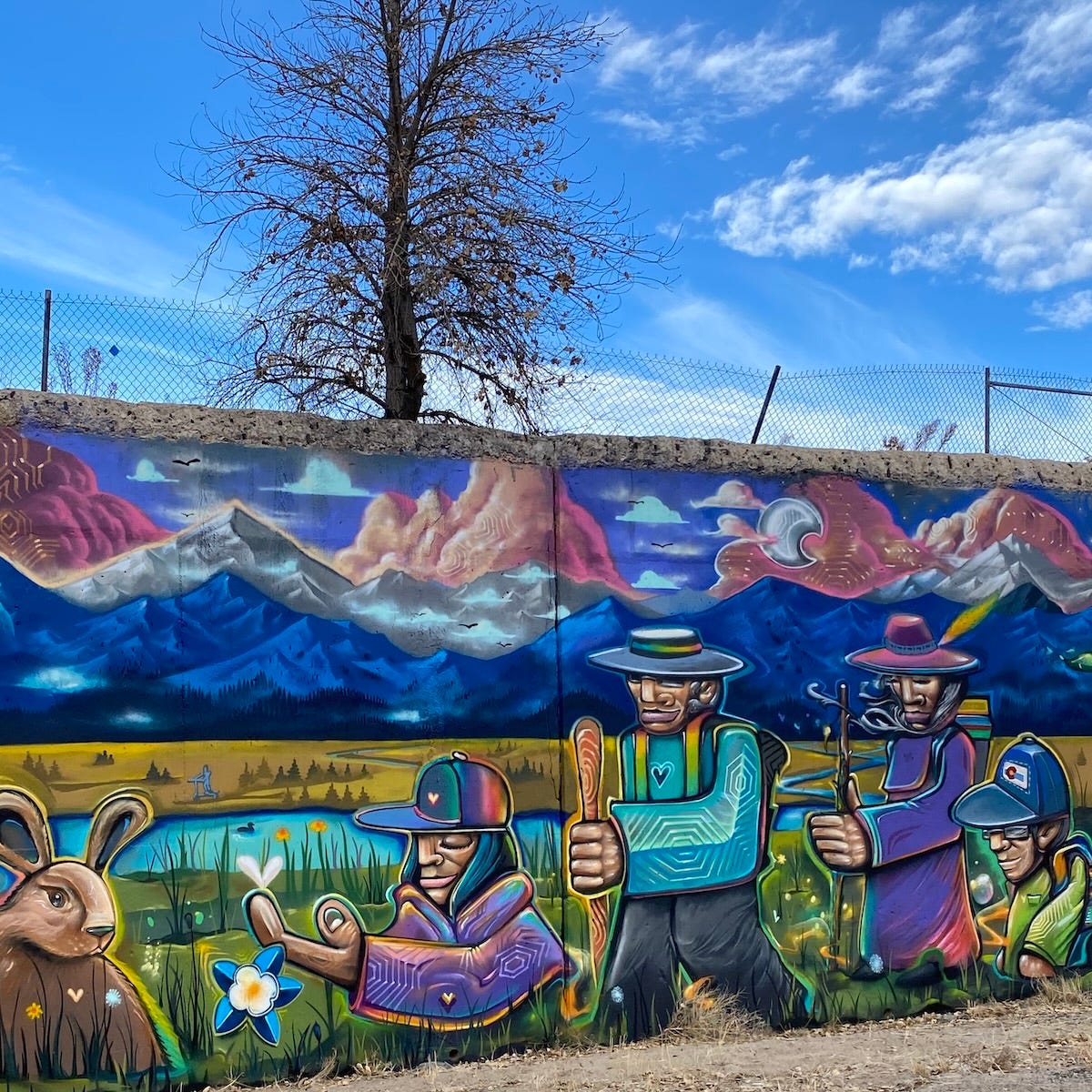 A mural painted on a wall depicting people, wildlife, flowers and mountains. Above the wall a tree grows in front of a decripit chain link fence.