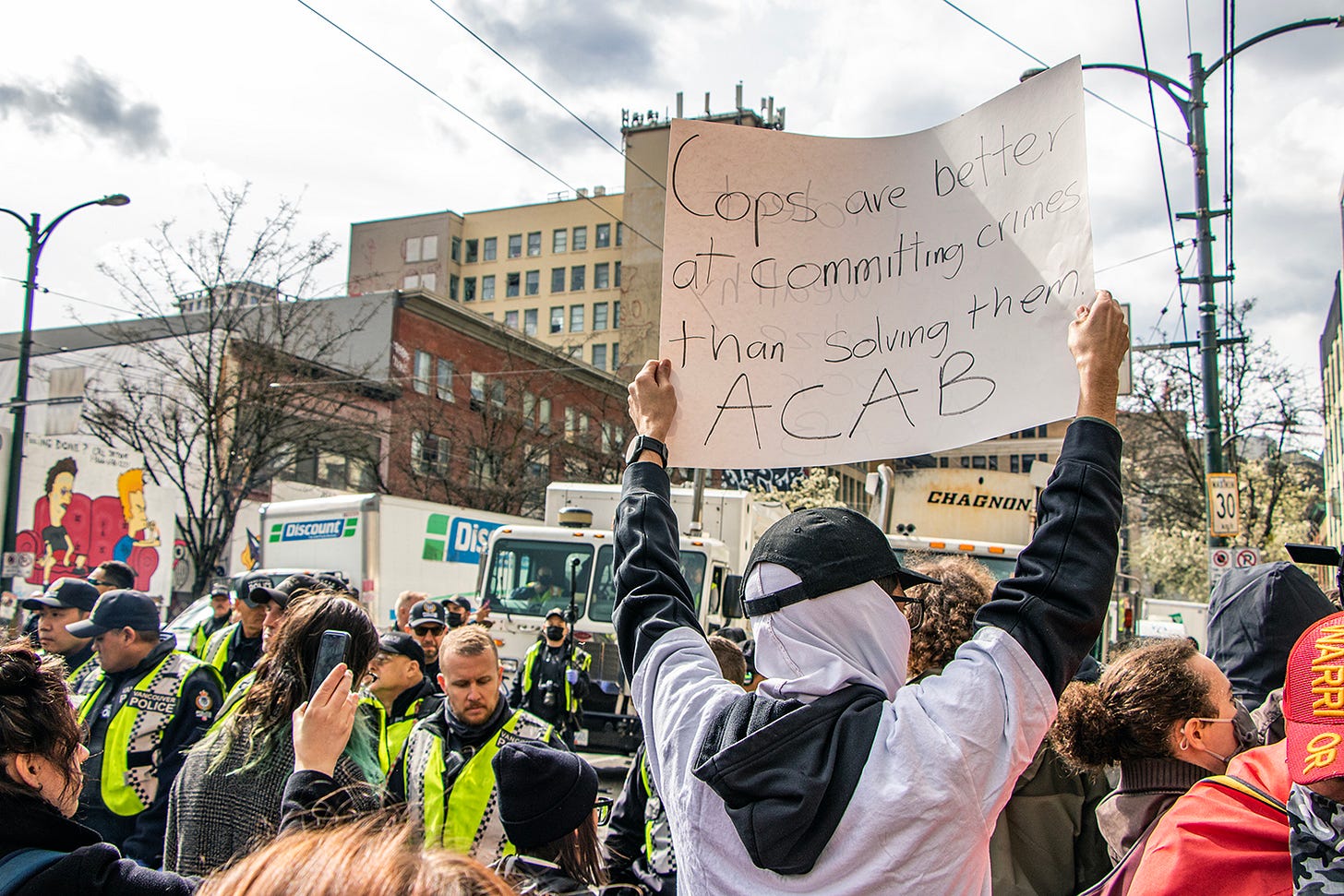 a person holds up a sign saying "cops are better at committing crimes than they are at solving them" at the front of a crowd, where they are lined up facing a wall of VPD officers. a number of box trucks and garbage trucks are behind the police line